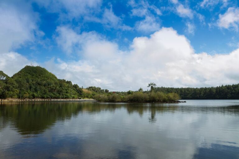 A serene view of the Grand Bassin Sacred Lake under a bright blue sky with scattered clouds. The lake is surrounded by lush greenery, including a small island with trees, and a hill covered in dense foliage on the left side.