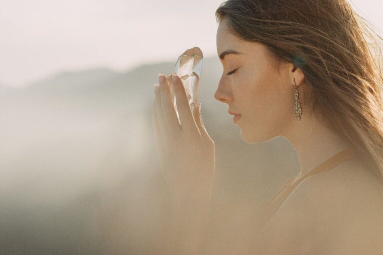 The image shows a serene, soft-focus scene of a woman holding a clear crystal up to her face, with her eyes closed in a peaceful, meditative pose. The sunlight gently illuminates her face and the crystal, casting a warm glow. The background is blurred with natural, earthy tones, enhancing the tranquil and reflective mood of the moment.