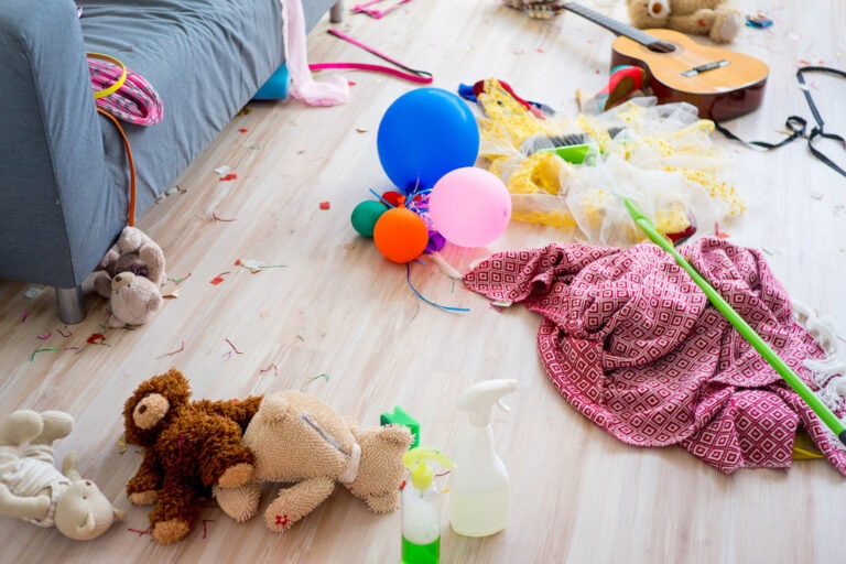 This image shows a cluttered living room with toys, balloons, confetti, and party decorations scattered across the floor. A guitar, teddy bears, a broom, and cleaning supplies are also visible, suggesting that the room is in need of tidying up after a celebration or event. The scene reflects a fun, chaotic aftermath, likely from a children's party or playful gathering.
