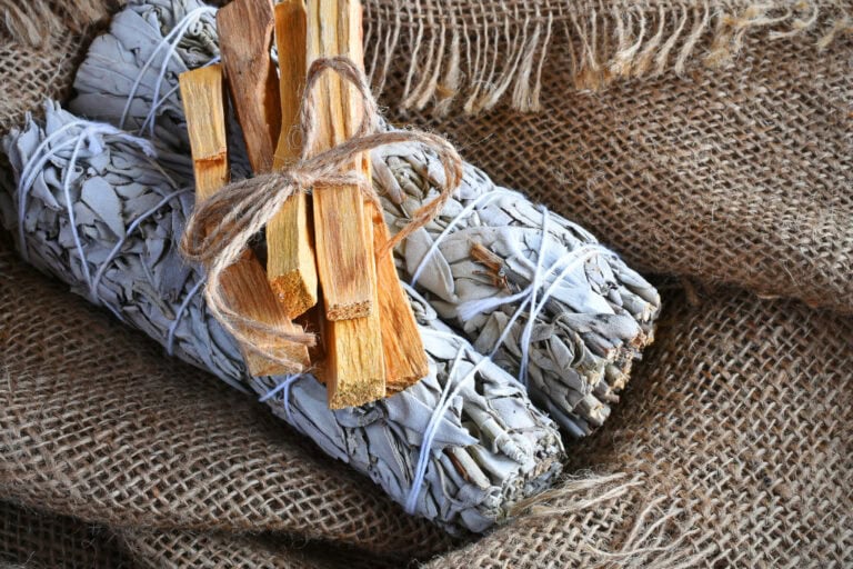 This image displays two sage bundles wrapped in white string, with a small bundle of Palo Santo wood tied on top with twine. The items rest on a textured burlap fabric, creating a rustic and earthy feel. These are commonly used for smudging and spiritual cleansing ceremonies.